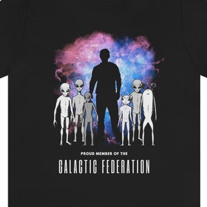 以下が含まれることがあります： 銀河を背景に、ピンクとブルーの色合いで、グレーの宇宙人のグループが人物のシルエットの前に立っているグラフィックがプリントされた黒いTシャツ。グラフィックの下には「Proud Member of the Galactic Federation」というテキストがプリントされています。