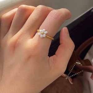 Puede incluir: Un anillo de flor de margarita blanca hecho con pequeñas cuentas blancas y doradas.