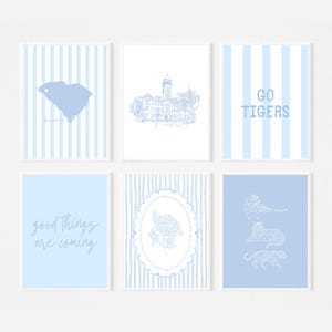 May include: Six framed art prints with a light blue and white color scheme. The prints feature a South Carolina map, a building sketch, the words "GO TIGERS", the phrase "good things are coming", a floral design, and tiger illustrations.