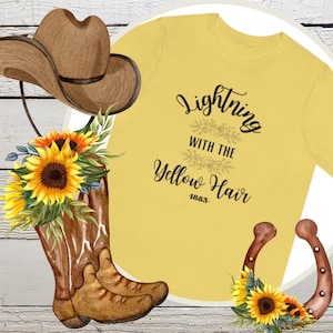 Könnte beinhalten: Gelbes T-Shirt mit dem Text "Lightning with the Yellow Hair -1883-." Das Shirt ist von einem Cowboyhut, Cowboystiefeln und Sonnenblumen umgeben.
