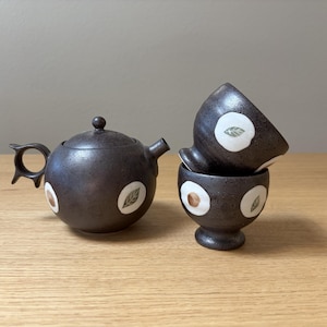 May include: A dark brown speckled ceramic teapot with a unique ring-shaped handle and two matching footed cups. Each piece features white circular accents containing small green leaf illustrations, creating a cohesive set for serving tea.