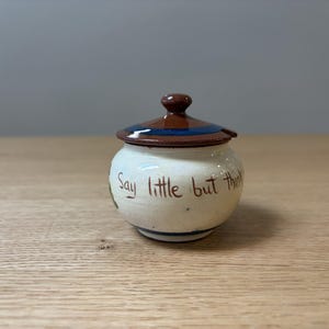 May include: A small, round, cream-colored ceramic jar with a matching lid. The lid features a dark brown top and a blue band. The body of the jar is inscribed with the brown text "Say little but think" and includes a small green painted detail.