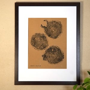 May include: Framed art print featuring three black tree ring illustrations on a tan background. The print is matted in white and framed in a dark brown wooden frame. The text "HAND MAPLE" is printed at the bottom.