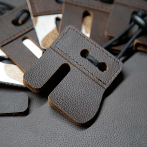 May include: Close-up of several brown leather accessories with a U-shaped design and a small hole. Each piece has a black cord attached. The background is a textured, dark gray surface.