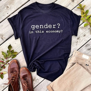 May include: A dark blue t-shirt with the text "gender? in this economy?" printed in white on the front. The shirt is laid out on a wooden surface with a pair of brown leather boots and a pair of khaki pants.