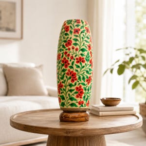 May include: A cylindrical vase with a cream-colored base featuring a hand-painted pattern of vibrant red flowers and green leafy vines. The vase is supported by a circular, polished dark brown wooden base, creating a distinct contrast in color.