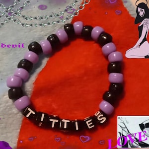 May include: A beaded bracelet with black and lavender beads spelling out the word "TITTIES". The bracelet is set against a red background with decorative images of devils and angels.