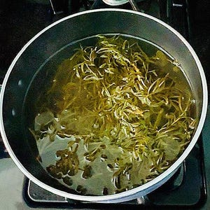 Puede incluir: Una olla de acero inoxidable llena de agua y hierbas verdes, probablemente para preparar t&eacute; o un remedio herbal. El agua es de color amarillo verdoso claro, y las hierbas son de un verde vibrante. La olla est&aacute; sobre una encimera negra.