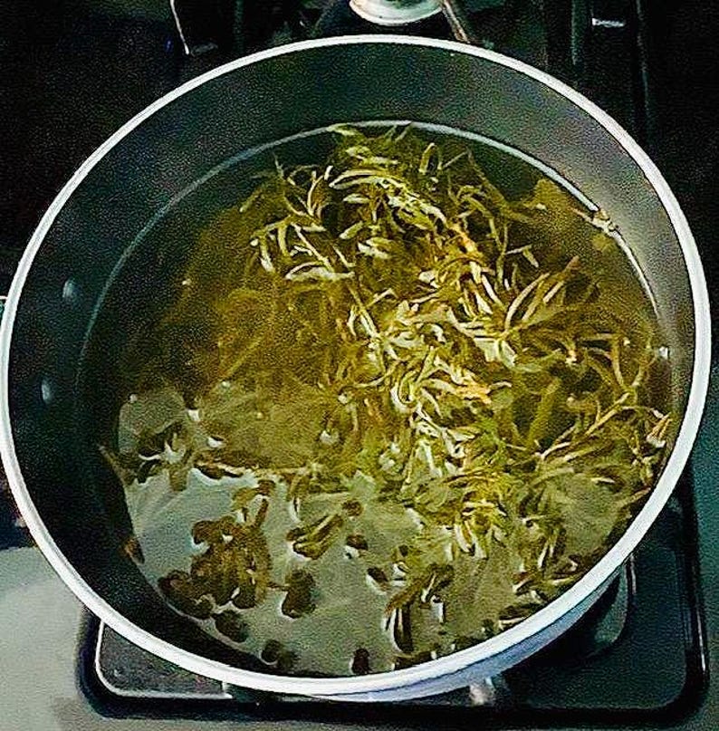 Puede incluir: Una olla de acero inoxidable llena de agua y hierbas verdes, probablemente para preparar t&eacute; o un remedio herbal. El agua es de color amarillo verdoso claro, y las hierbas son de un verde vibrante. La olla est&aacute; sobre una encimera negra.