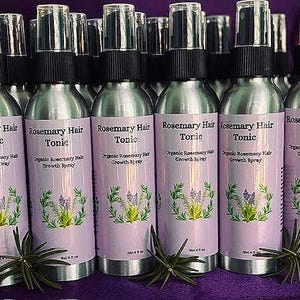 Puede incluir: Una colecci&oacute;n de botellas de spray plateadas con tapas negras, etiquetadas como "Rosemary Hair Tonic". Cada botella tiene una etiqueta lila claro con texto y una ilustraci&oacute;n bot&aacute;nica. Las botellas est&aacute;n sobre una superficie morada.