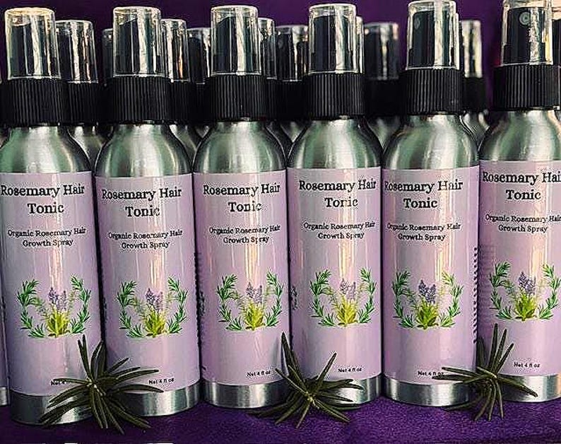 Puede incluir: Una colecci&oacute;n de botellas de spray plateadas con tapas negras, etiquetadas como "Rosemary Hair Tonic". Cada botella tiene una etiqueta lila claro con texto y una ilustraci&oacute;n bot&aacute;nica. Las botellas est&aacute;n sobre una superficie morada.