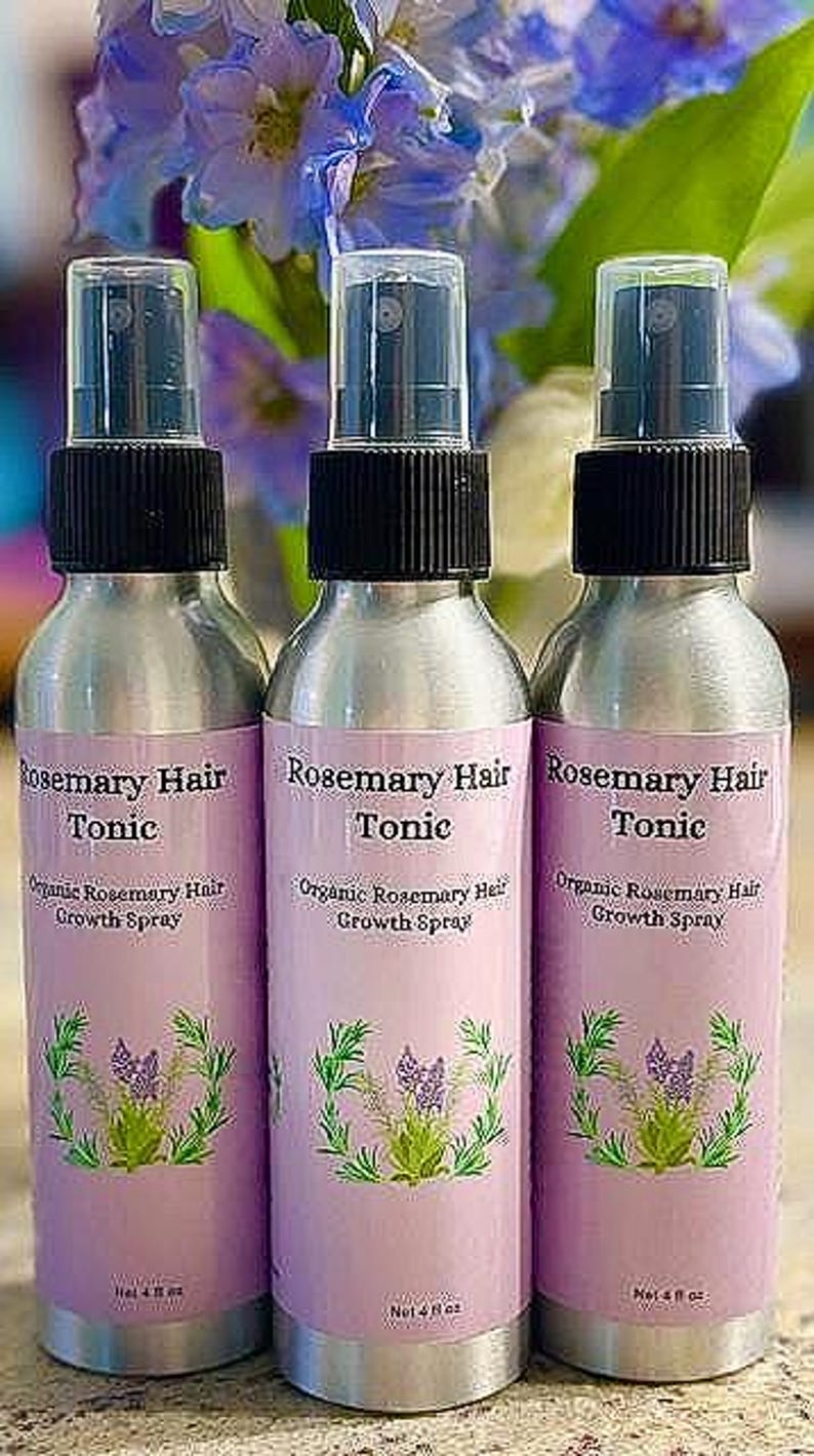 Puede incluir: Tres botellas de spray plateadas de Rosemary Hair Tonic con tapas negras y etiquetas rosas. Las etiquetas presentan el texto "Rosemary Hair Tonic" y "Organic Rosemary Hair Growth Spray" con una ilustraci&oacute;n floral. Las botellas son de 118 ml.