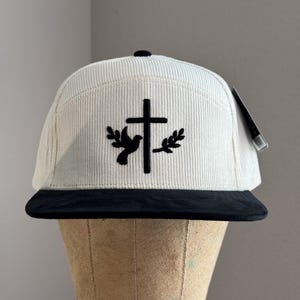 May include: A white corduroy baseball cap with a black brim. The cap features a black embroidered design of a dove with an olive branch on each side of a cross.
