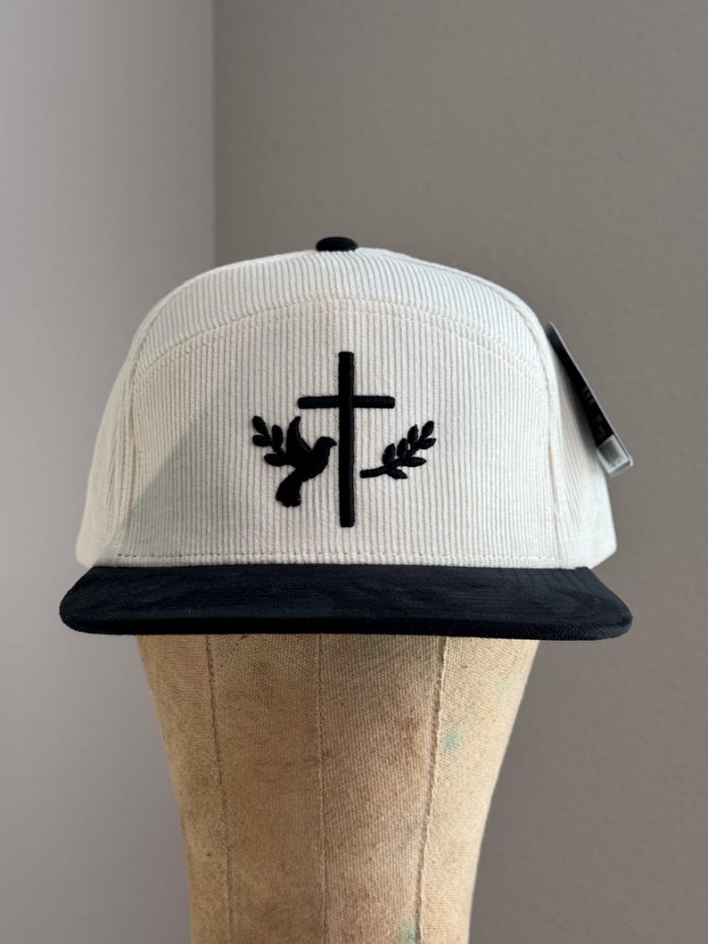 May include: A white corduroy baseball cap with a black brim. The cap features a black embroidered design of a dove with an olive branch on each side of a cross.