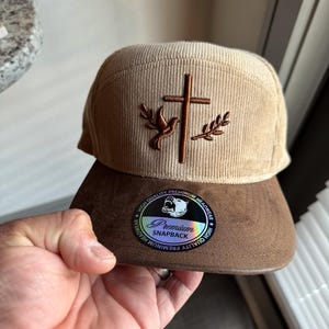 May include: A tan corduroy baseball cap with a brown suede bill. The cap features an embroidered cross with a dove and olive branches. The bill has a sticker that reads "Premium Snapback" with a bear logo.