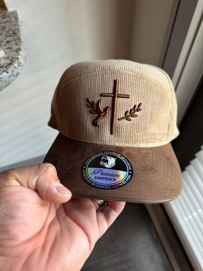 May include: A tan corduroy baseball cap with a brown suede bill. The cap features an embroidered cross with a dove and olive branches. The bill has a sticker that reads "Premium Snapback" with a bear logo.