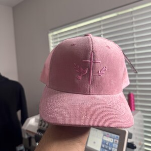 May include: A pink corduroy baseball cap with a mesh back. The cap features an embroidered design of a dove and cross with olive branches.