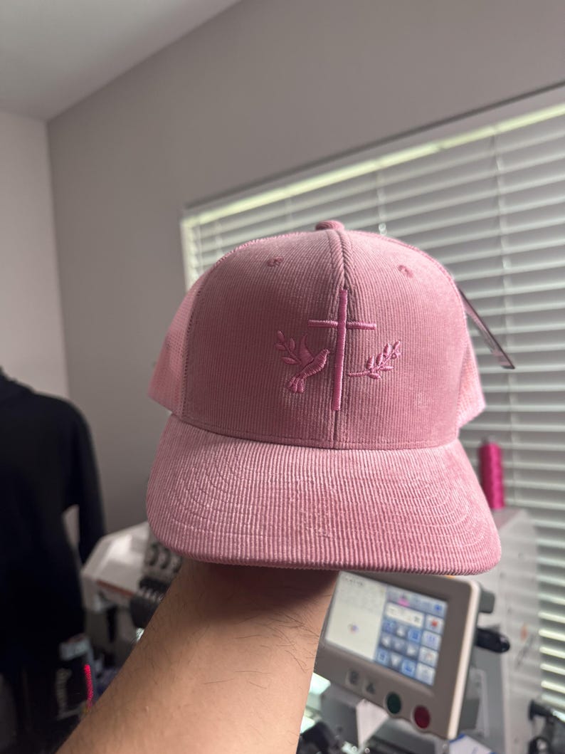 May include: A pink corduroy baseball cap with a mesh back. The cap features an embroidered design of a dove and cross with olive branches.