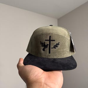May include: A green corduroy baseball cap with a black embroidered cross and dove design. The cap has a black suede brim.