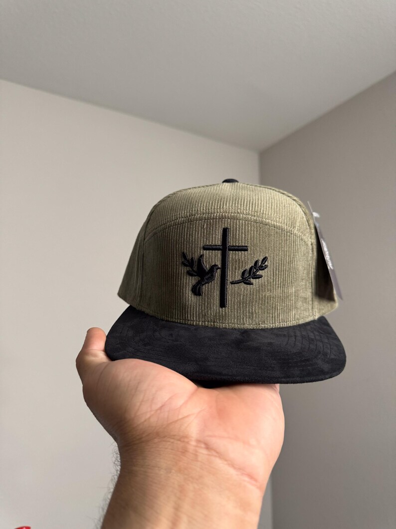 May include: A green corduroy baseball cap with a black embroidered cross and dove design. The cap has a black suede brim.