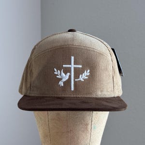 May include: A brown corduroy baseball cap with a brown suede brim. The cap features a white embroidered cross with a dove and olive branches on either side.