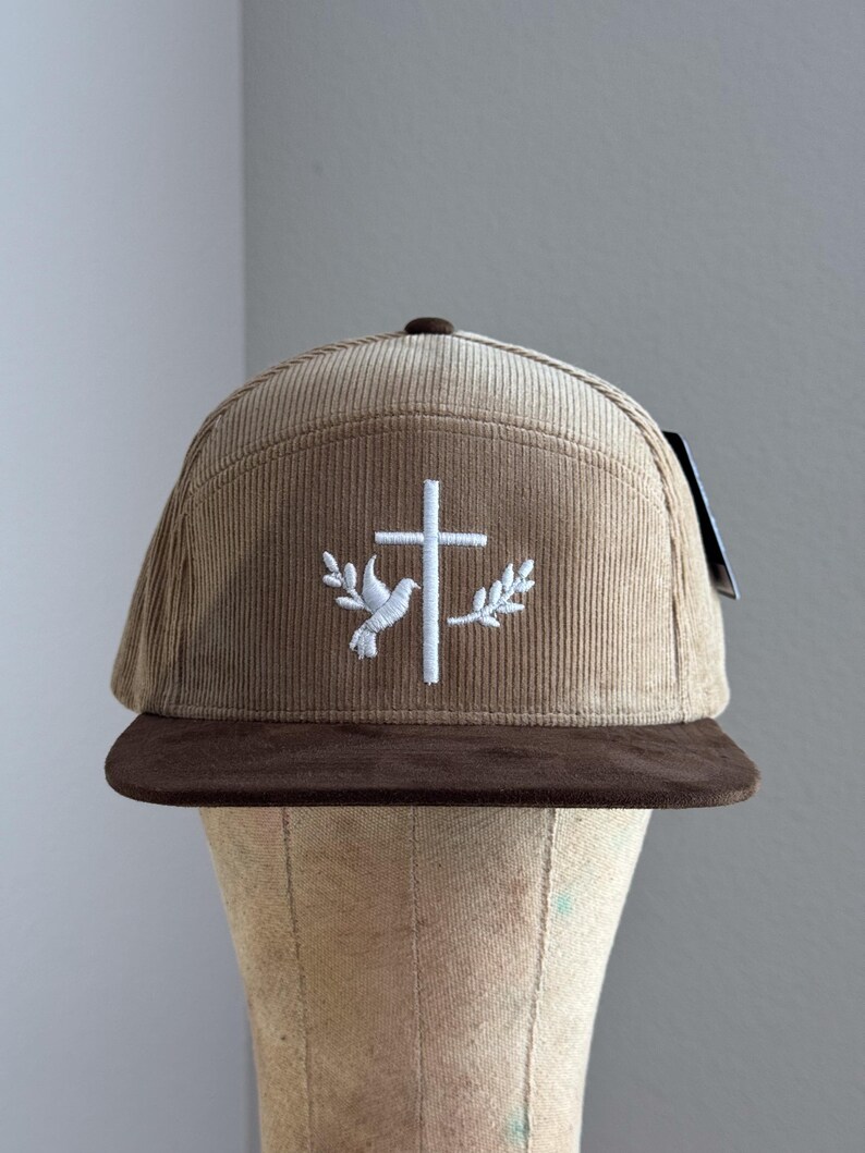 May include: A brown corduroy baseball cap with a brown suede brim. The cap features a white embroidered cross with a dove and olive branches on either side.