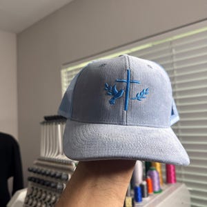 May include: A light blue corduroy trucker hat with a blue embroidered dove and cross design. The hat has a mesh back panel.