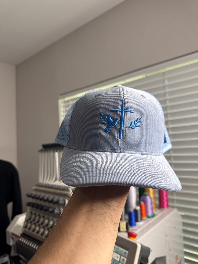 May include: A light blue corduroy trucker hat with a blue embroidered dove and cross design. The hat has a mesh back panel.