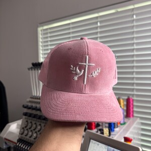 May include: A pink corduroy baseball cap with a white embroidered dove and cross design.