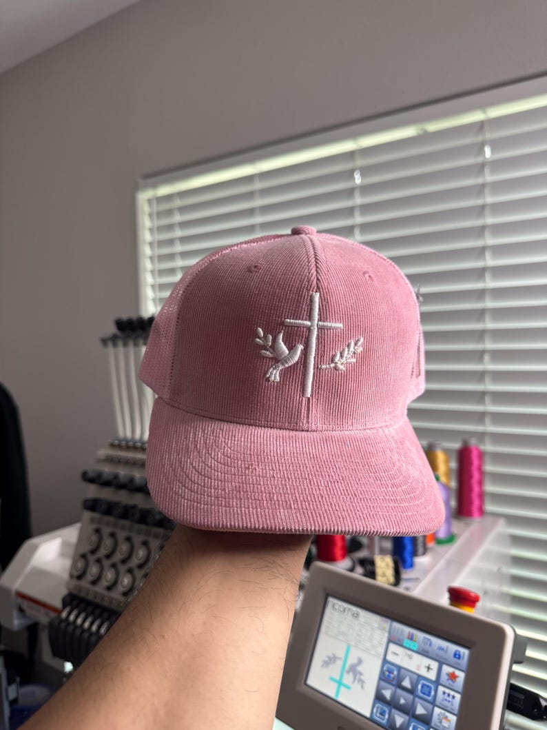 May include: A pink corduroy baseball cap with a white embroidered dove and cross design.
