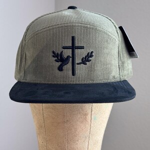 May include: A green corduroy baseball cap with a black brim. The cap features an embroidered design of a dove with an olive branch on each side of a cross.