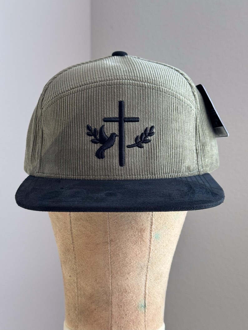 May include: A green corduroy baseball cap with a black brim. The cap features an embroidered design of a dove with an olive branch on each side of a cross.