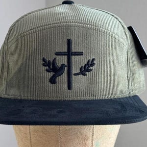 May include: A green corduroy baseball cap with a black brim. The cap features an embroidered design of a dove and a cross.