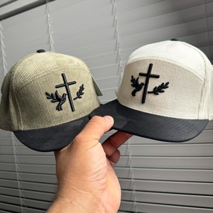 May include: Two corduroy baseball caps with a black cross and a white dove with olive branches on the front. One cap is beige and the other is green.