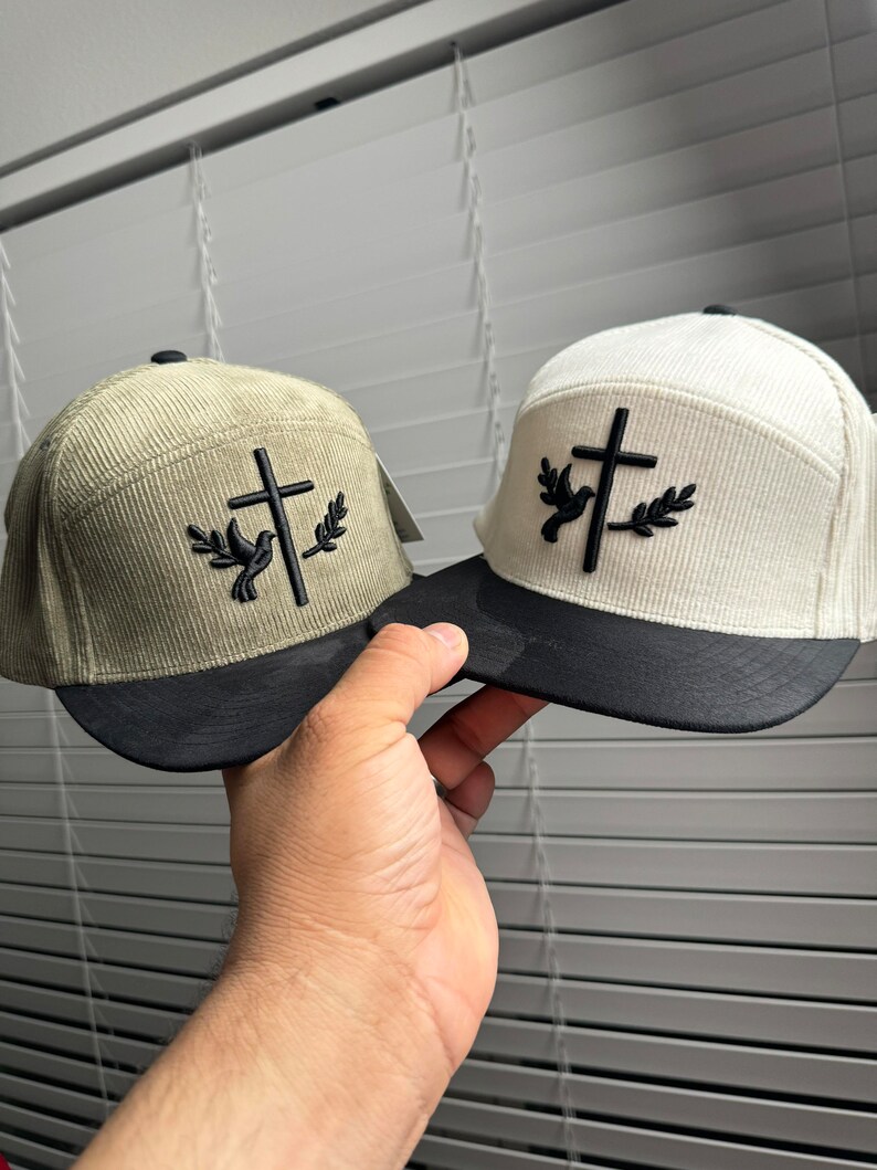 May include: Two corduroy baseball caps with a black cross and a white dove with olive branches on the front. One cap is beige and the other is green.