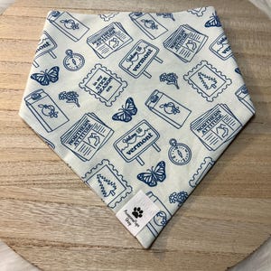 Può includere: Un bandana per cani azzurro e bianco con un motivo ripetuto di francobolli, farfalle, bussole e cartelli con la scritta "Welcome to Vermont" e "Northern Attitude". Il bandana ha un'etichetta con la scritta "Aussome Pups Shop".