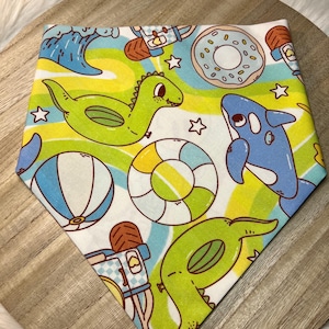 May include: A white bandana with a colorful summer-themed print. The design includes cartoon images of a green dinosaur float, a blue orca float, a beach ball, a donut float, and a wave. The background has yellow and blue stripes.