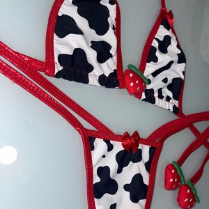 May include: A two-piece bikini set featuring a cow print design with black spots on a white background. The edges are trimmed in red, and small red bows adorn the straps. Includes strawberry accents.