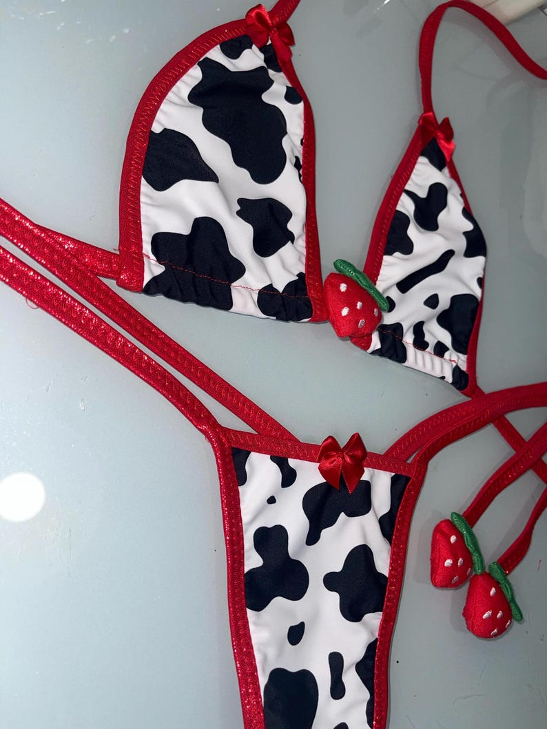 May include: A two-piece bikini set featuring a cow print design with black spots on a white background. The edges are trimmed in red, and small red bows adorn the straps. Includes strawberry accents.