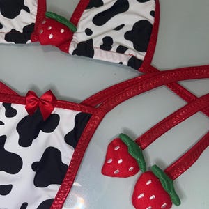 May include: A two-piece bikini set with a cow print pattern in black and white. The edges are trimmed with red fabric. The top features red strawberry accents. The bottom has a red bow.