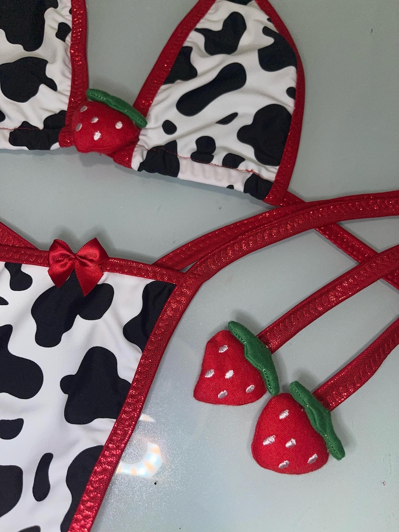 May include: A two-piece bikini set with a cow print pattern in black and white. The edges are trimmed with red fabric. The top features red strawberry accents. The bottom has a red bow.
