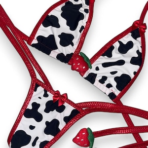 May include: A two-piece lingerie set featuring a cow print design with black spots on a white background. The set includes a triangle top and a thong bottom, both trimmed in red. Small red bows and strawberry accents add detail.