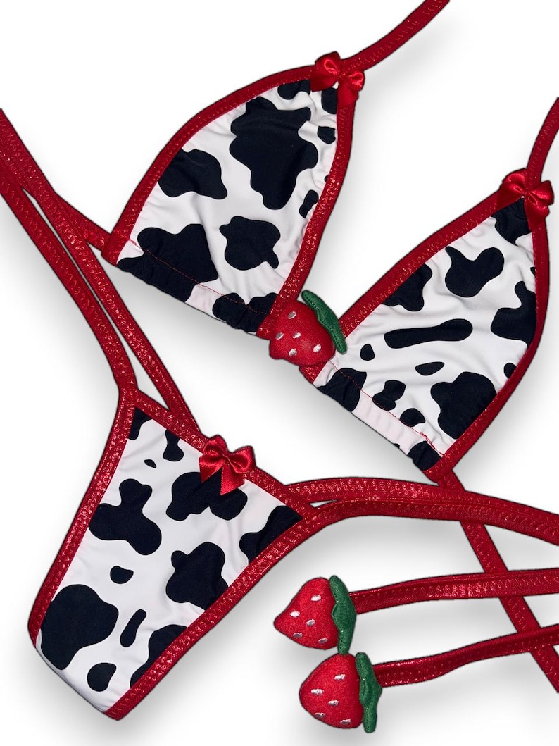 May include: A two-piece lingerie set featuring a cow print design with black spots on a white background. The set includes a triangle top and a thong bottom, both trimmed in red. Small red bows and strawberry accents add detail.