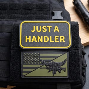 Puede incluir: Un chaleco táctico negro con dos parches rectangulares. El parche superior tiene fondo negro, borde amarillo y el texto amarillo JUST A HANDLER. El parche inferior muestra un diseño de bandera estadounidense verde con un emblema alado central.