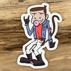 May include: A cartoon illustration of a monkey wearing a blue denim jacket, white pants, and black boots. The monkey is holding a peace sign with one hand and has a bubble of pink gum in its mouth. The monkey has brown fur and a red shirt underneath the jacket.