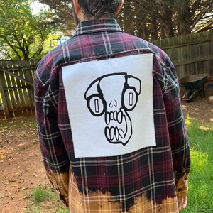 May include: A red and black plaid flannel shirt with a white patch on the back. The patch features a black and white illustration of a skull with large eyes.