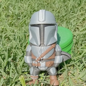 K&ouml;nnte beinhalten: Eine 3D-gedruckte Mandalorianer-Figur im Chibi-Stil. Die Figur ist haupts&auml;chlich grau mit braunen und schwarzen Akzenten, einschlie&szlig;lich Helm, R&uuml;stung und Blaster. Sie steht auf einer gr&uuml;nen Grasfl&auml;che.