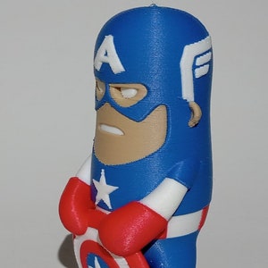 May include: A blue and red 3D printed figurine of Captain America holding his shield. The figurine is in a standing pose with his arms crossed and his shield in front of him. The shield is white with a red star in the center.