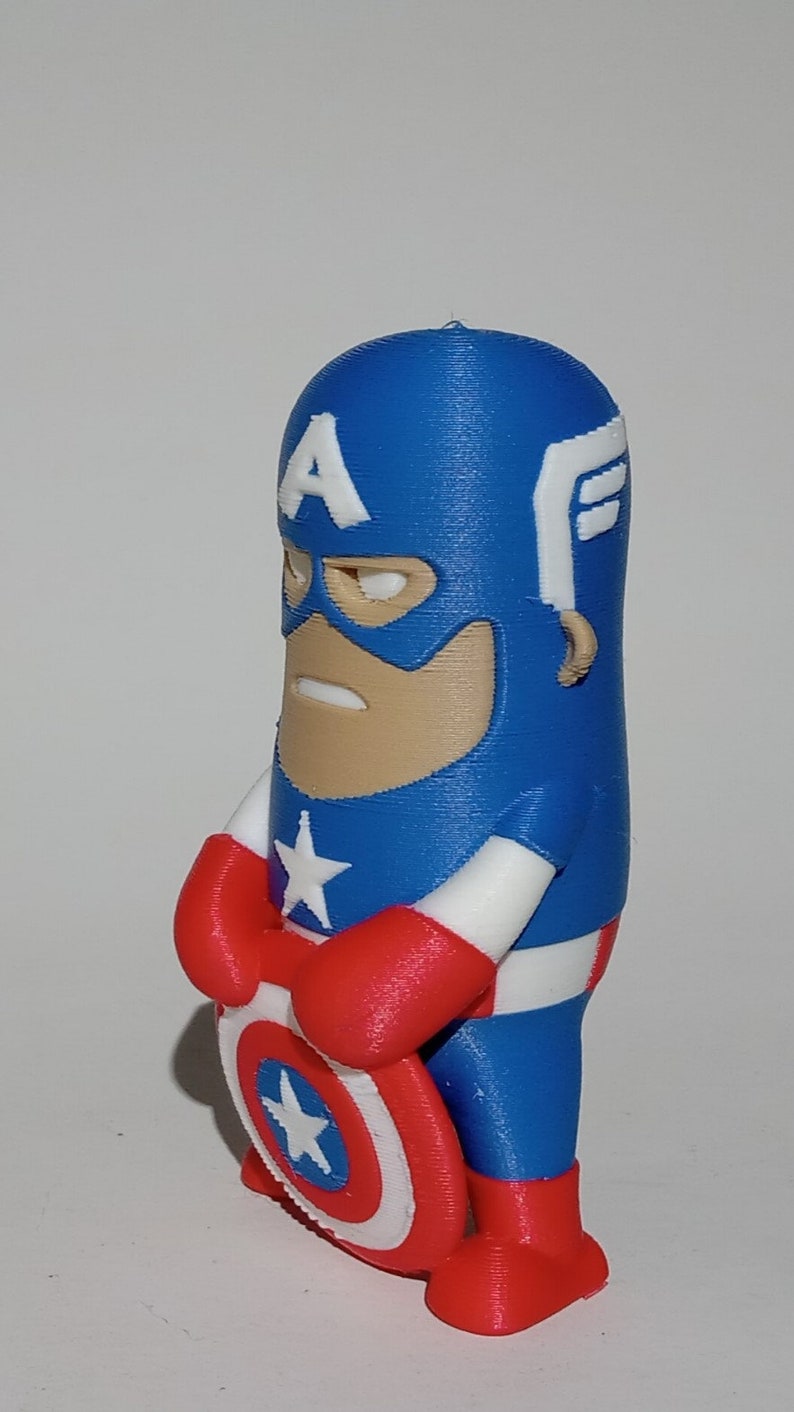 May include: A blue and red 3D printed figurine of Captain America holding his shield. The figurine is in a standing pose with his arms crossed and his shield in front of him. The shield is white with a red star in the center.