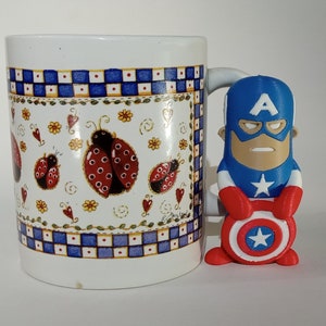 May include: A white ceramic mug with a blue and white checkered pattern and a ladybug design. A small red, white, and blue Captain America figurine is holding a red, white, and blue shield.